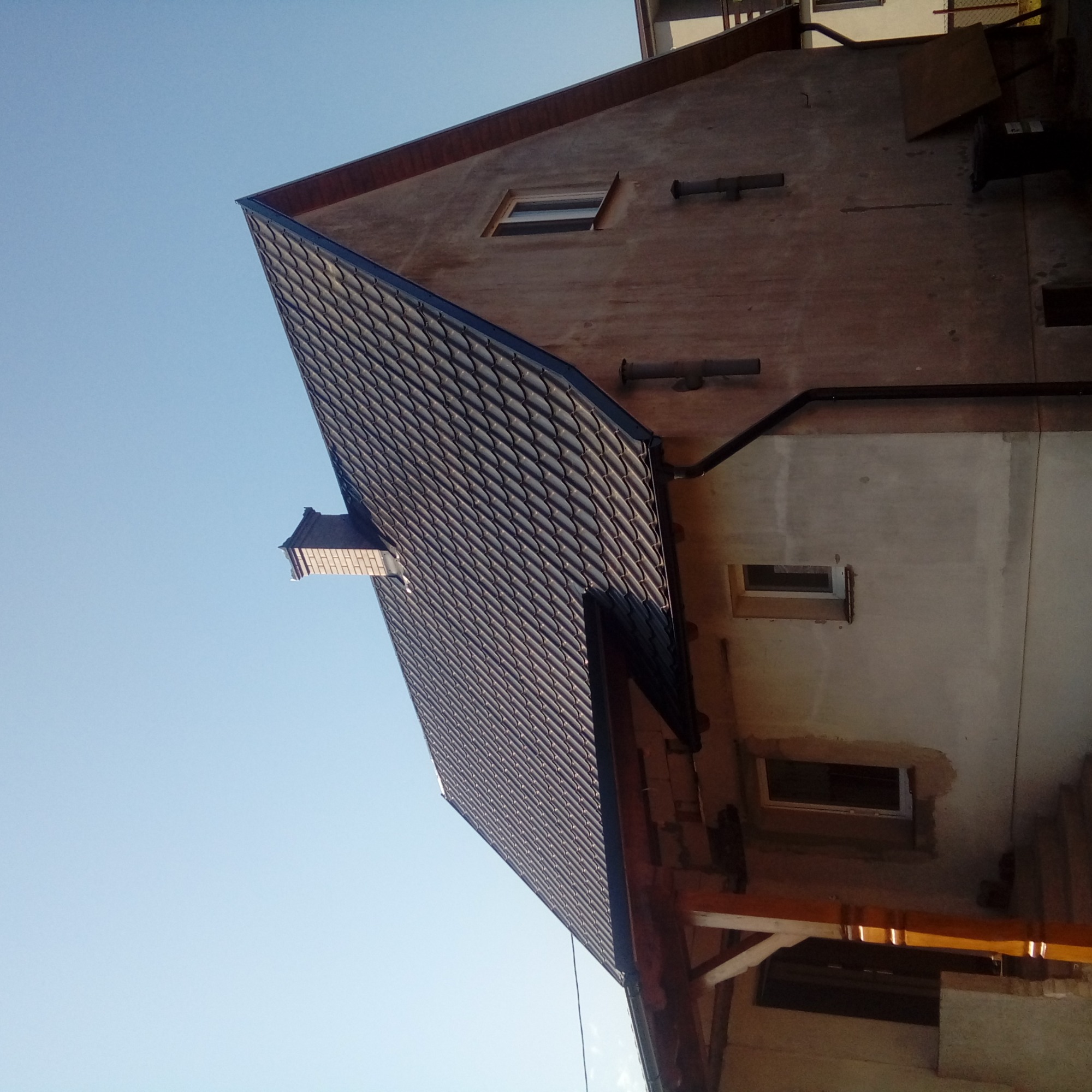 Nowy dach pokryty ciemną dachówką karpiówką, widoczny komin i orynnowanie, elewacja budynku w trakcie prac wykończeniowych.