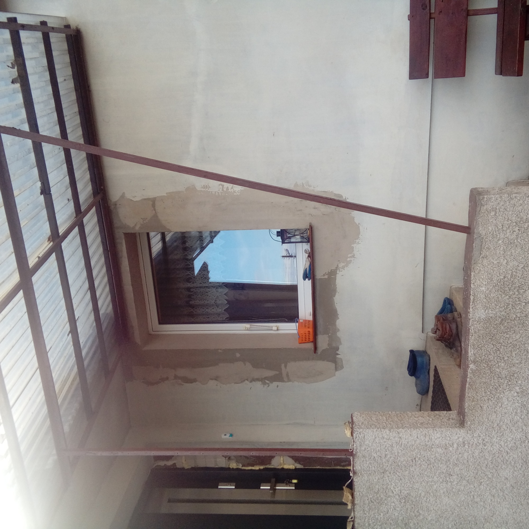 Ściana budynku z oknem po wymianie, widoczne ślady świeżego tynku, na parapecie robocze rękawice i pomarańczowy koszyk, na schodach robocze buty, zadaszenie z blachy trapezowej.
