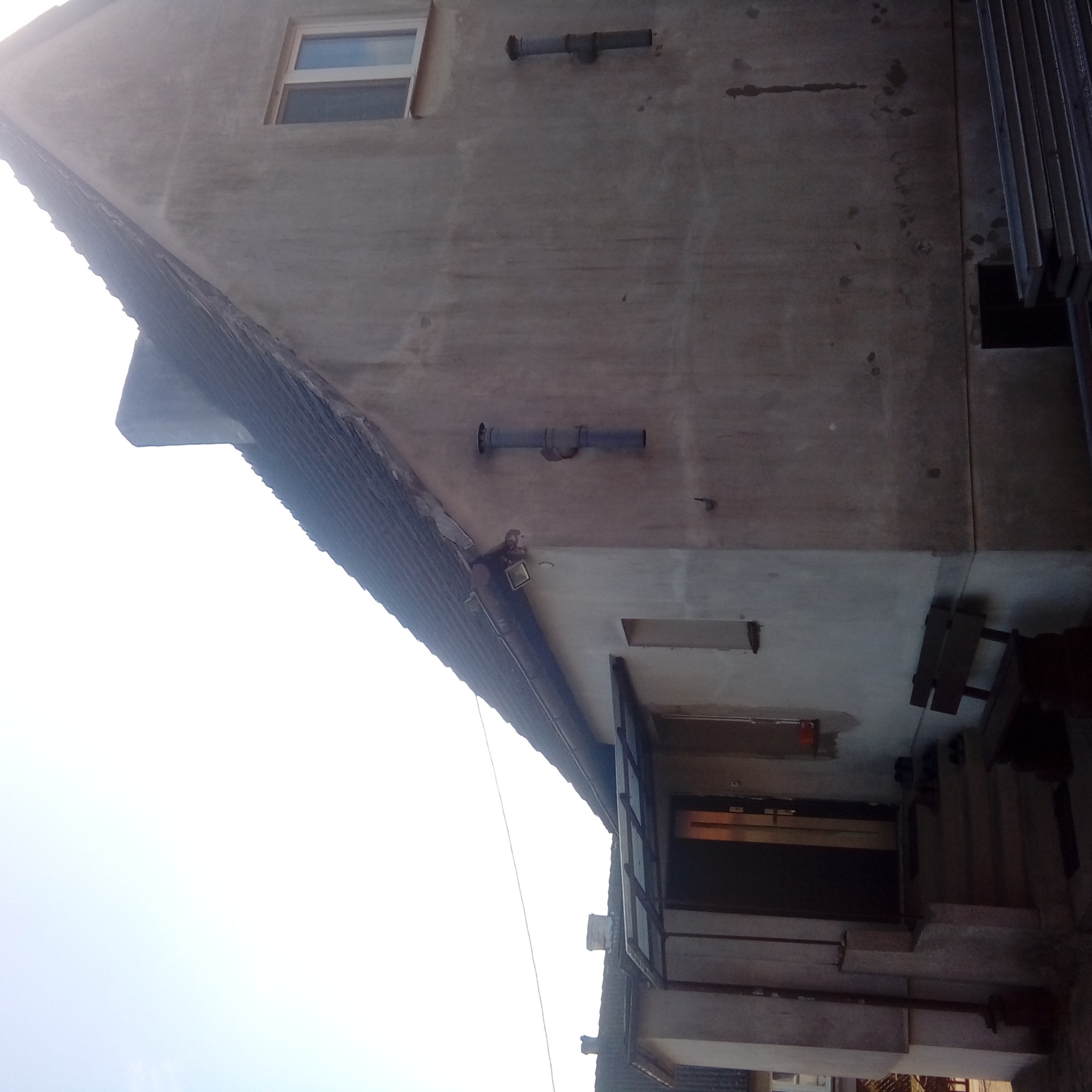 Niski kąt widzenia na szary budynek z widoczną częścią dachu z dachówką, rynnami i oknem, z elementami instalacji na elewacji.