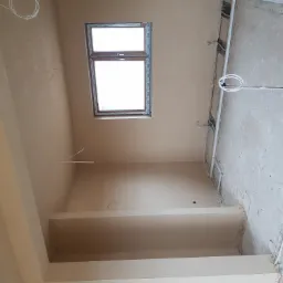 Wnętrze w trakcie prac, widoczne świeżo nałożone tynki na ścianach i suficie, instalacja elektryczna prowadzona w korytach, okno dachowe z drewnianą ramą.