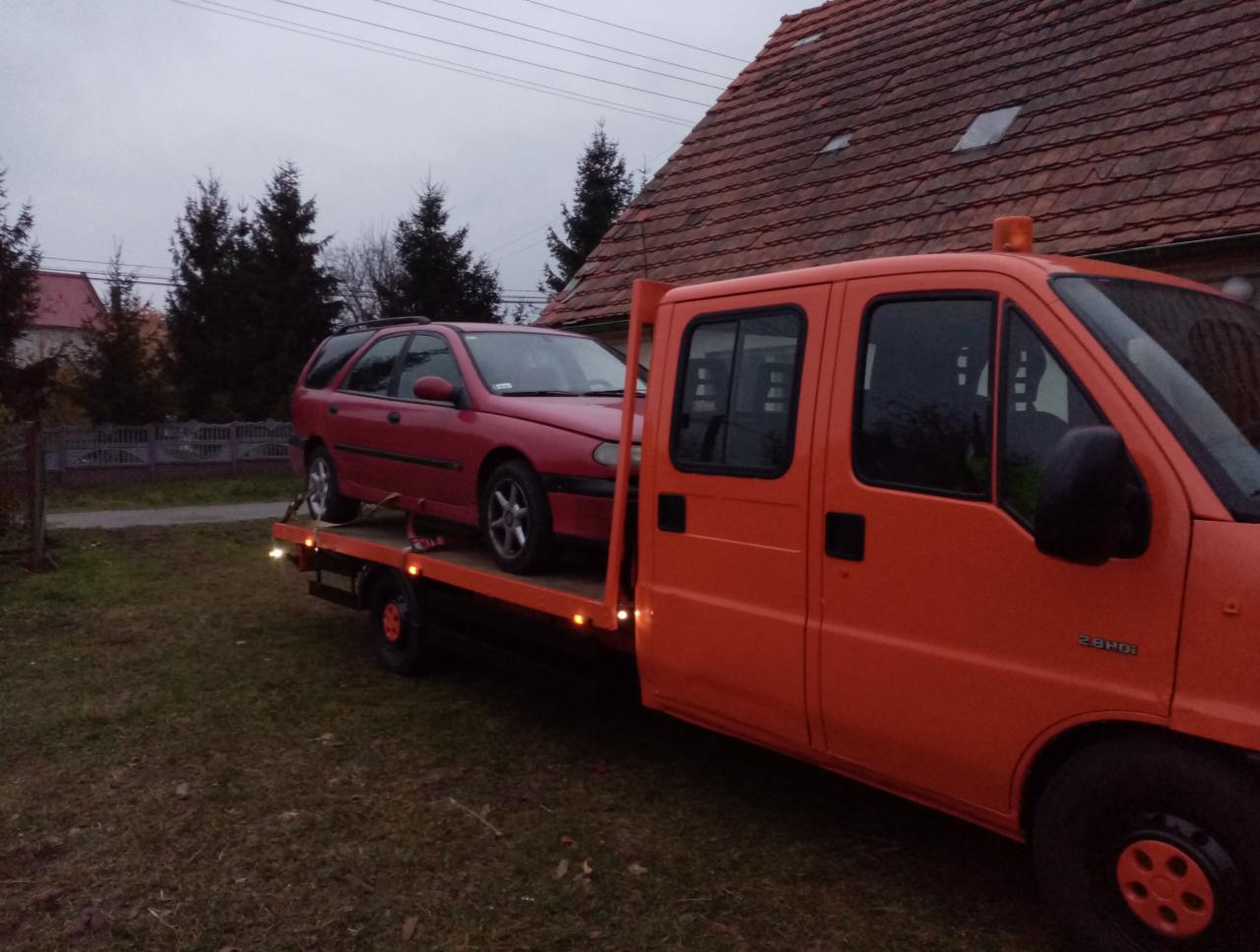 Pomarańczowy samochód pomocy drogowej z czerwonym samochodem osobowym na platformie, stojący na trawniku przed domem z czerwoną dachówką w pochmurny dzień.