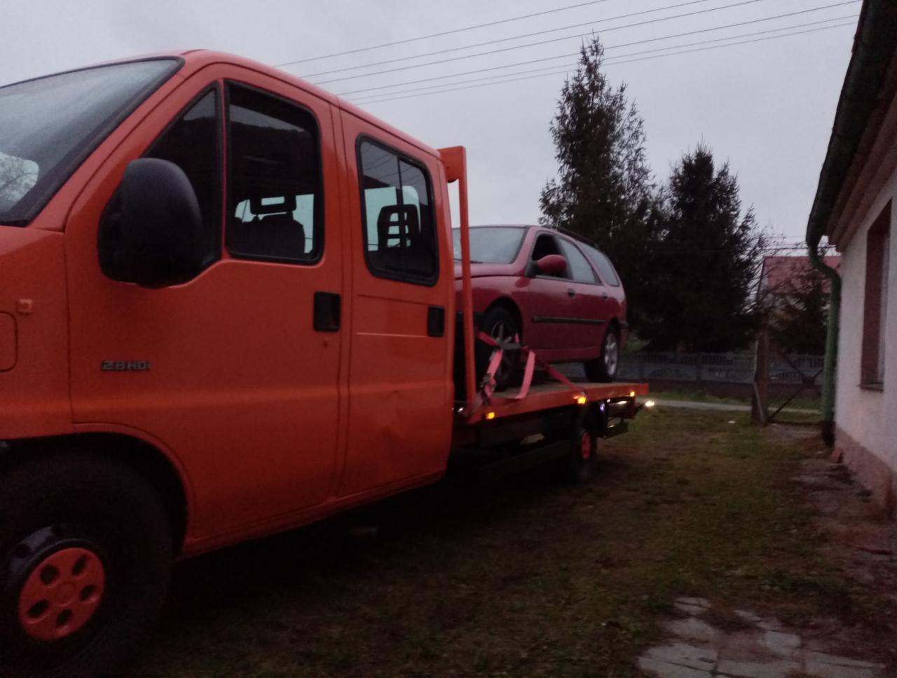 Pomarańczowa laweta z czerwonym samochodem osobowym na platformie, widok z boku na tle pochmurnego nieba i budynków.