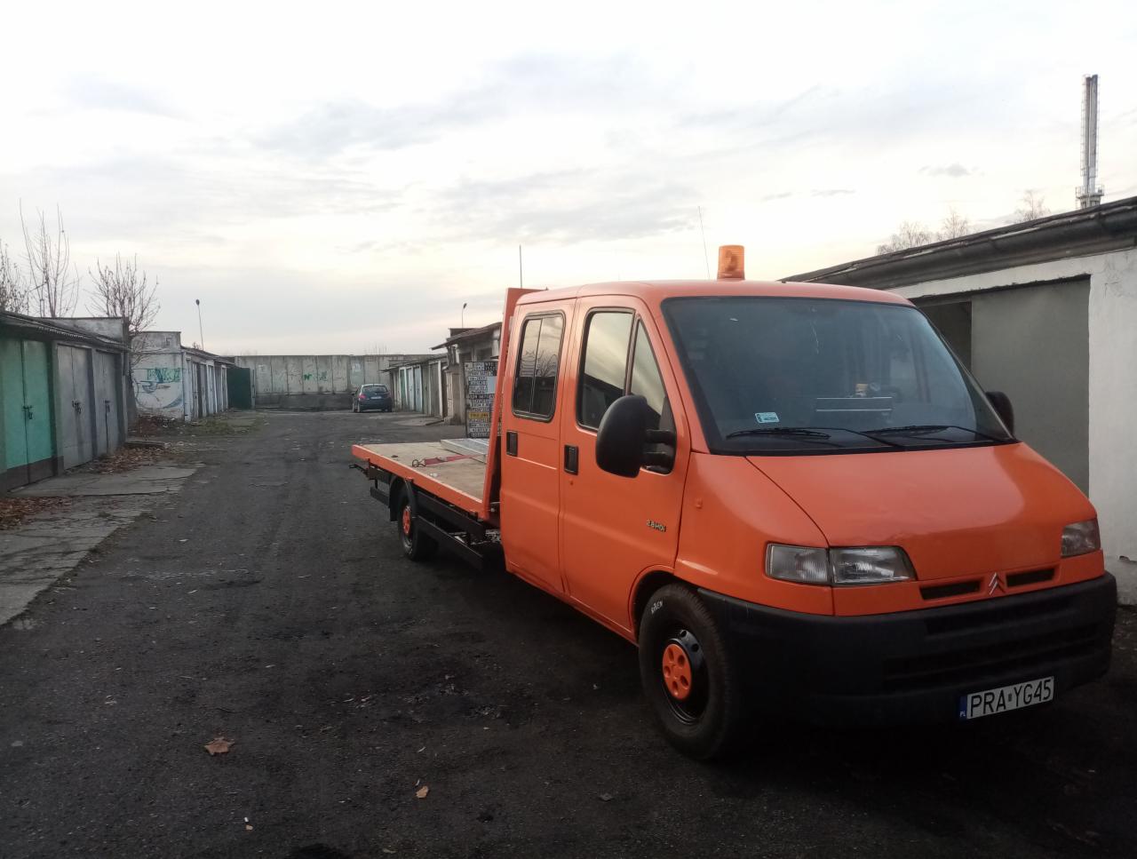 Pomarańczowy samochód z platformą transportową na parkingu, widoczne garaże w tle i tablica rejestracyjna PRA-YG45.