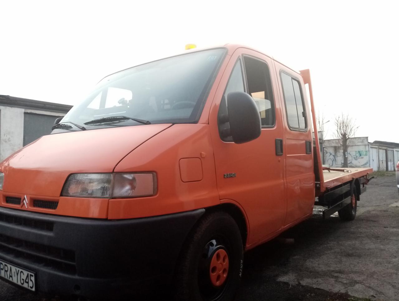 Pomarańczowy bus z platformą do przewozu pojazdów, widoczny z przodu i z boku na tle zabudowań gospodarczych.