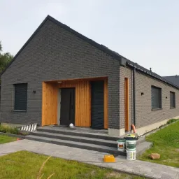 Nowoczesny dom z szarej cegły z drewnianym wejściem i schodami z kostki brukowej, widoczne puszki z farbą przy schodach.