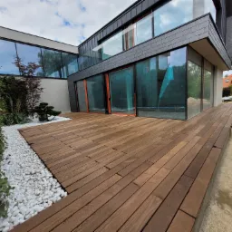 Nowoczesny dom z drewnianym tarasem, wykończonym białymi kamieniami i zielenią, widok na przeszkloną fasadę.