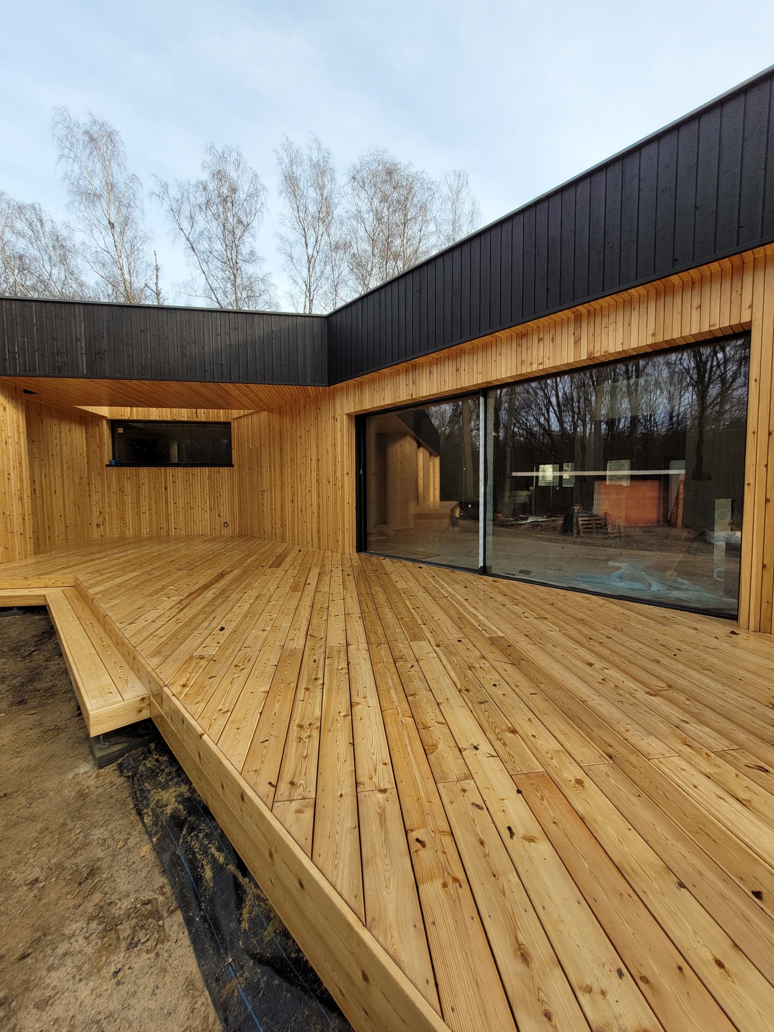 Nowoczesny taras z jasnego drewna, przylegający do budynku z czarną elewacją i dużymi oknami, widok na otaczający las.