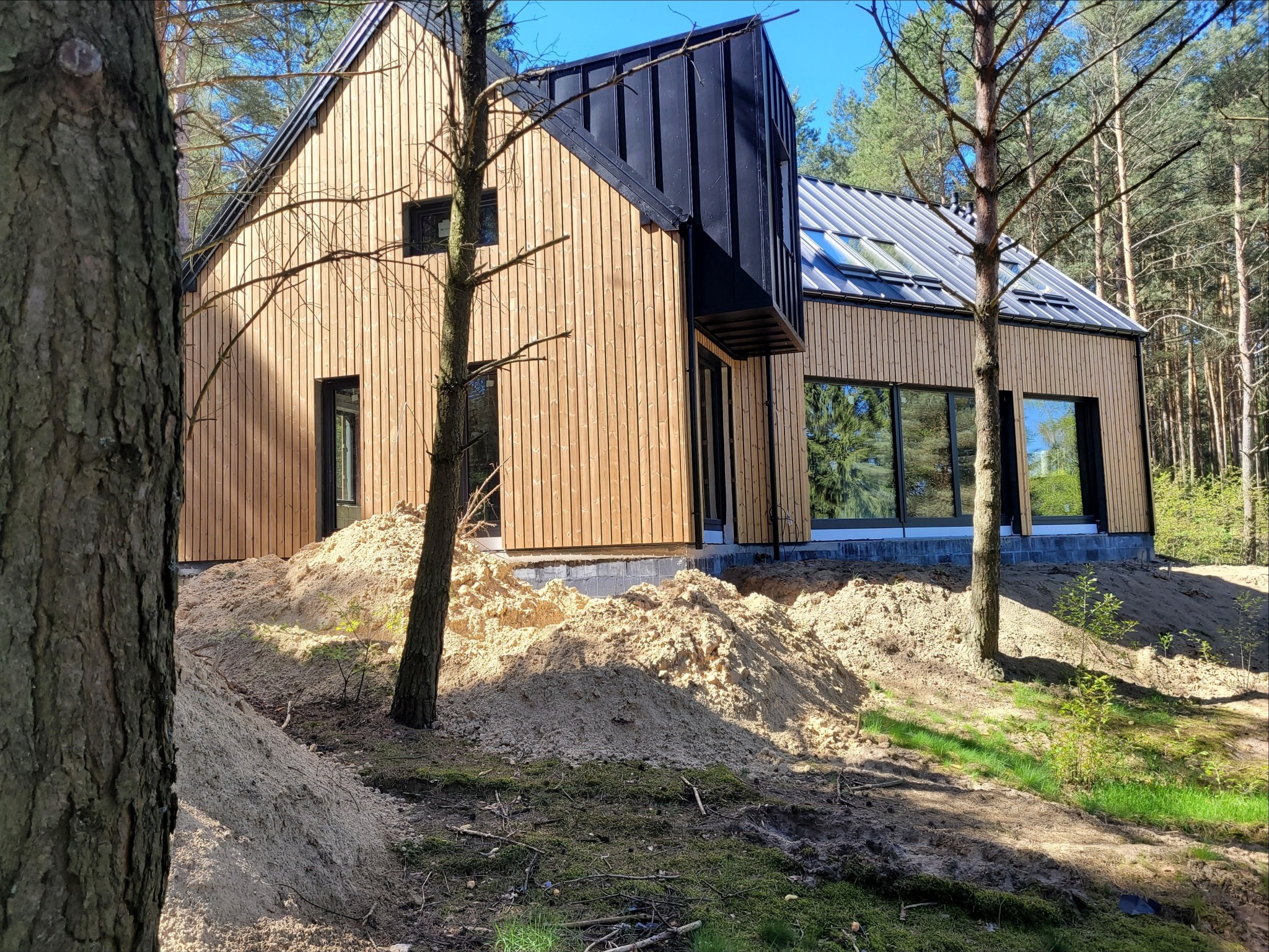 Nowoczesny dom z drewnianą elewacją i czarnym dachem, otoczony sosnowym lasem i usypaną ziemią.