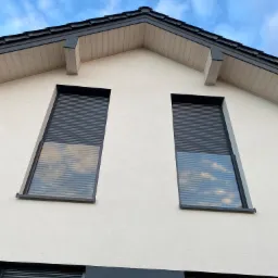 Elewacja domu z dwoma oknami wyposażonymi w częściowo opuszczone, grafitowe osłony okienne, odbijające błękitne niebo z chmurami. Dolna część elewacji również w kolorze grafitowym.