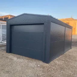 Nowy, grafitowy garaż blaszany z segmentową bramą garażową, ustawiony na żwirowym podłożu, widoczny w świetle popołudniowym.