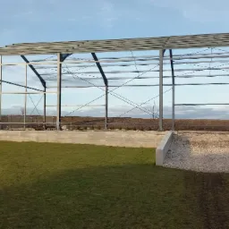 Stalowa konstrukcja hali w trakcie budowy, widoczna na tle zielonego trawnika i pola. Fundament betonowy z rampą wjazdową. Niebo częściowo zachmurzone.