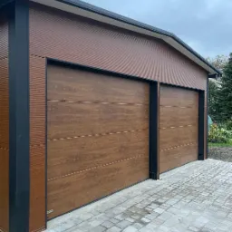Nowoczesny garaż blaszany z dwoma bramami segmentowymi o fakturze drewna, czarne obramowania, kostka brukowa, pochmurne niebo w tle.