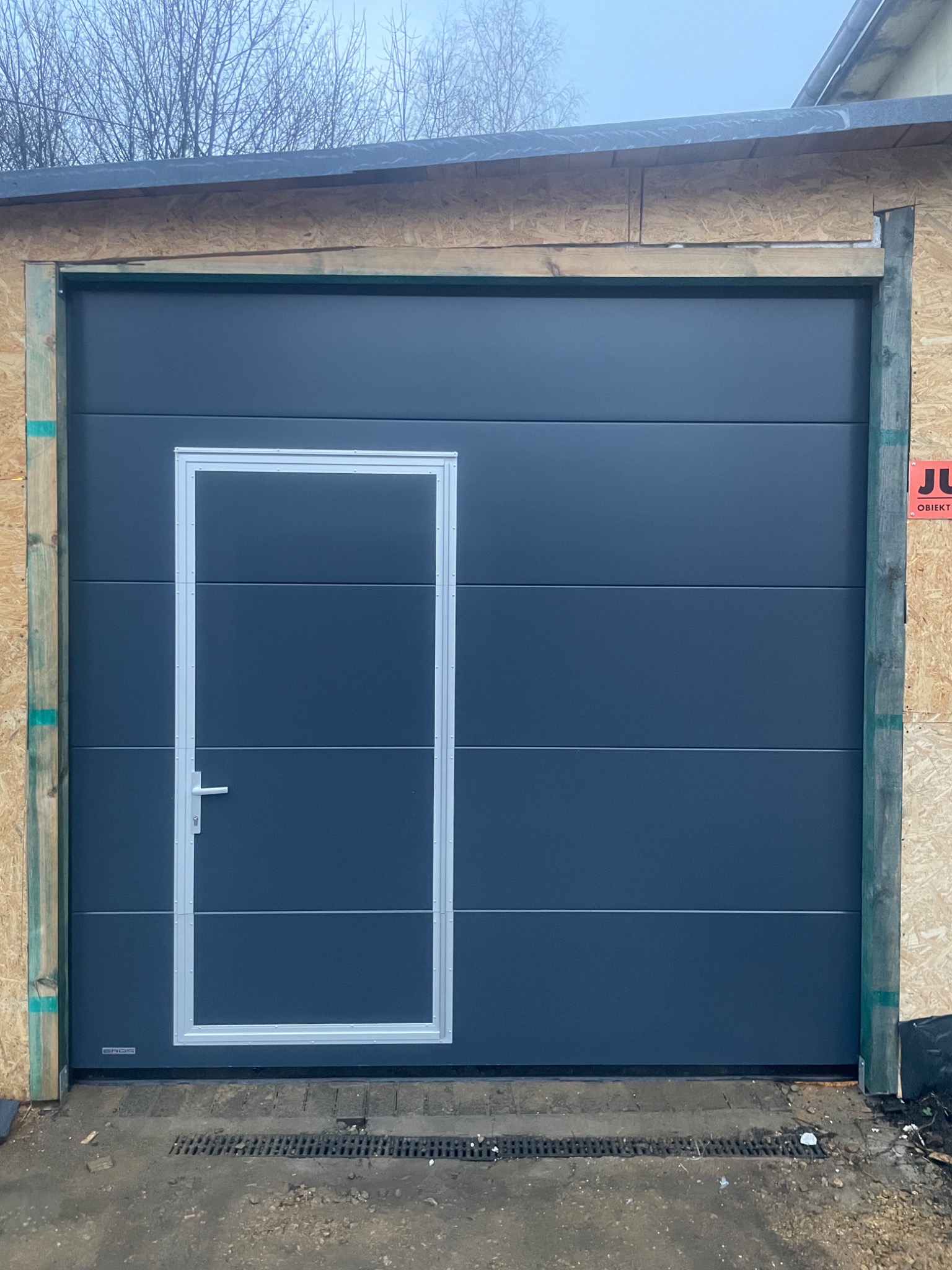 Szara segmentowa brama garażowa z drzwiami wejściowymi pośrodku, w trakcie montażu, widoczne drewniane elementy konstrukcyjne i tabliczka informacyjna.