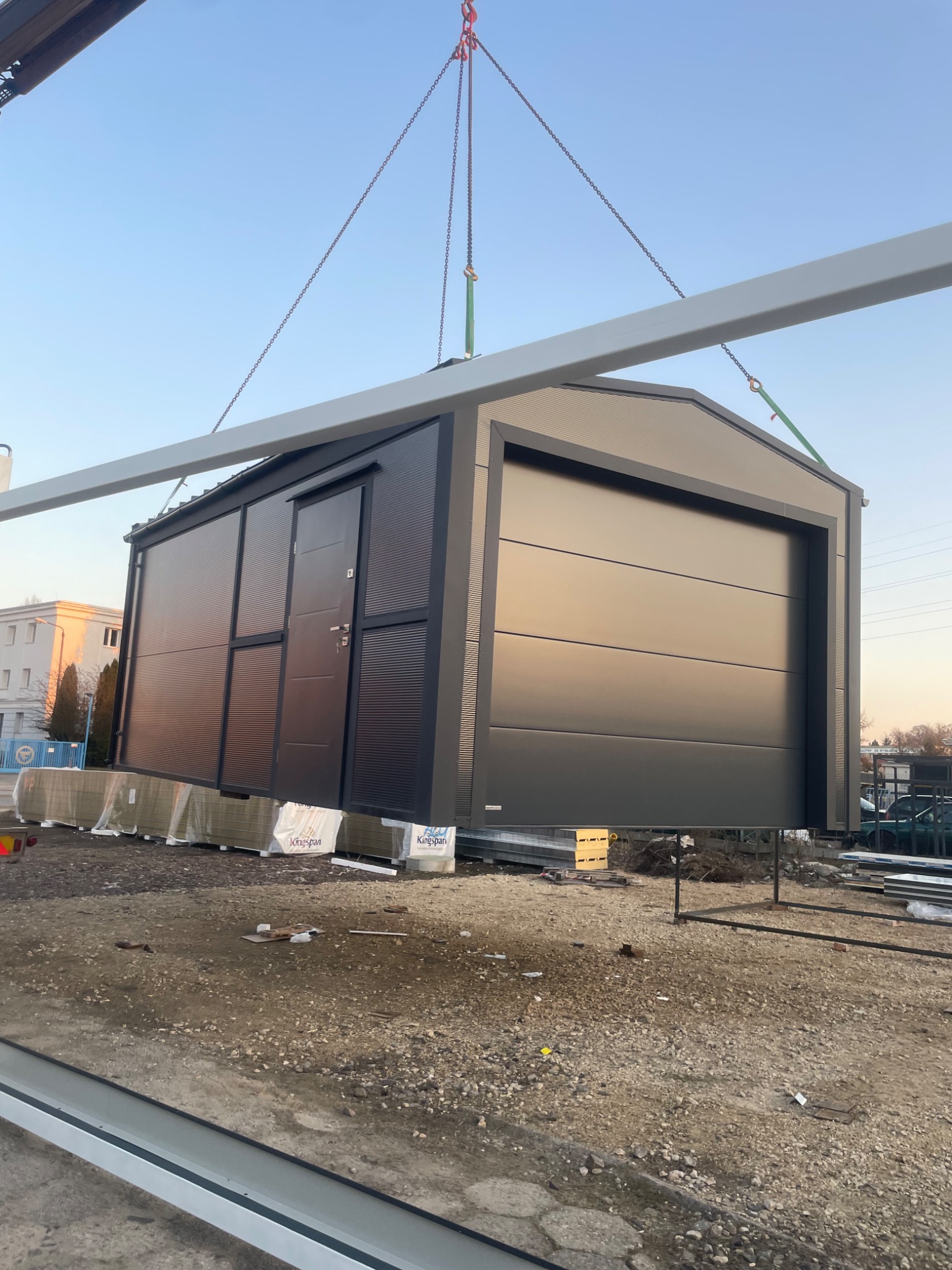 Czarny garaż z blachy trapezowej z bramą segmentową i drzwiami bocznymi, podnoszony dźwigiem na placu budowy.