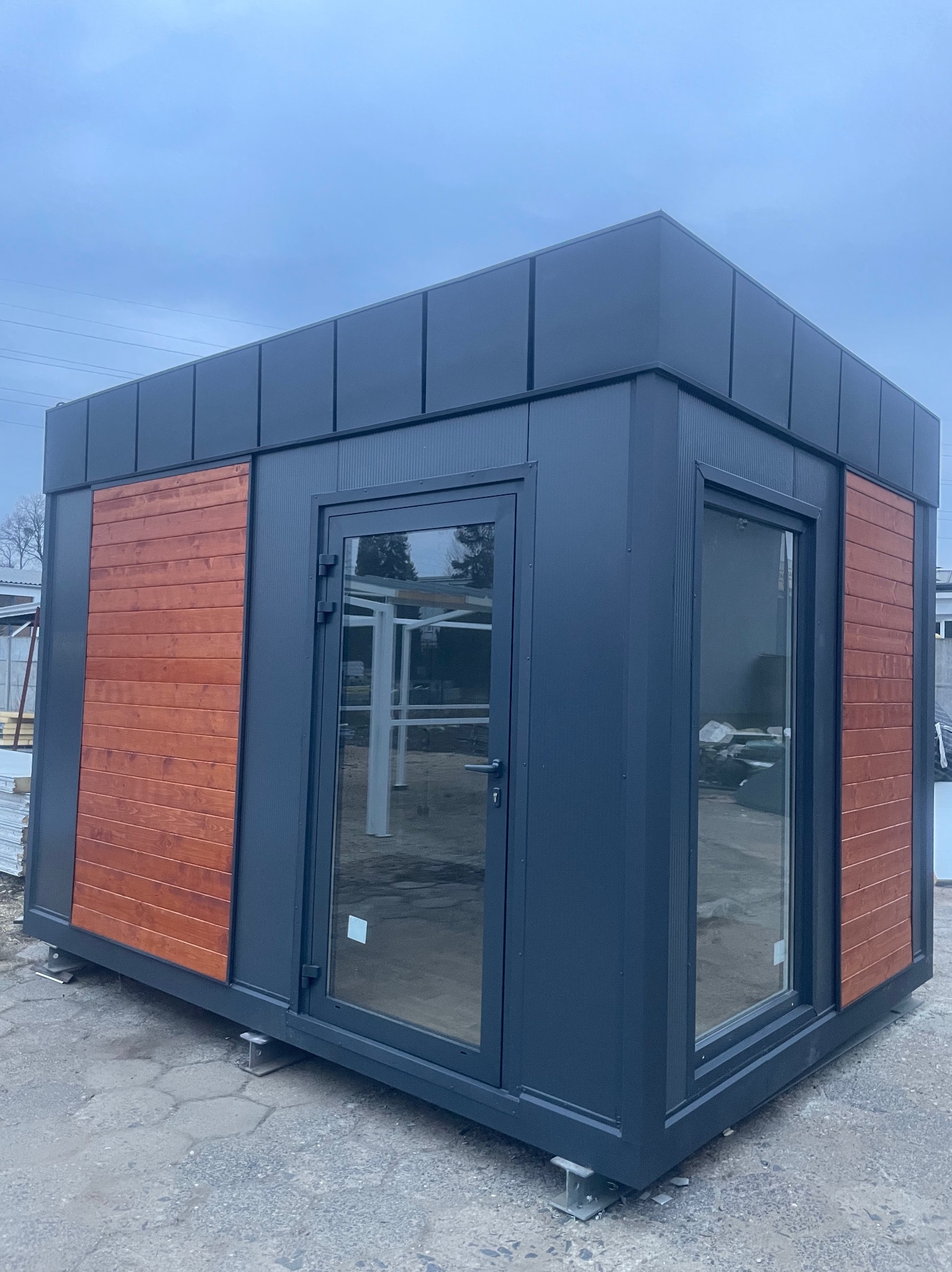 Nowoczesny, szary pawilon handlowy z drewnianymi panelami, przeszklonymi ścianami i drzwiami, ustawiony na betonowej nawierzchni w pochmurny dzień.