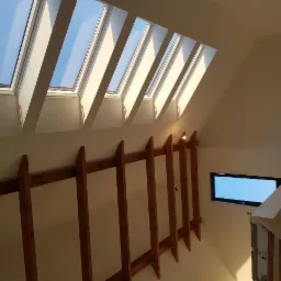 Poddasze z drewnianą konstrukcją pod sufitem, okna dachowe wpuszczające jasne światło dzienne i małe okno na ścianie, widok z góry na balustradę schodów.