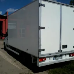 Tył białego samochodu dostawczego z zamkniętymi drzwiami, widoczna tablica rejestracyjna i fragment czerwonego budynku w tle.