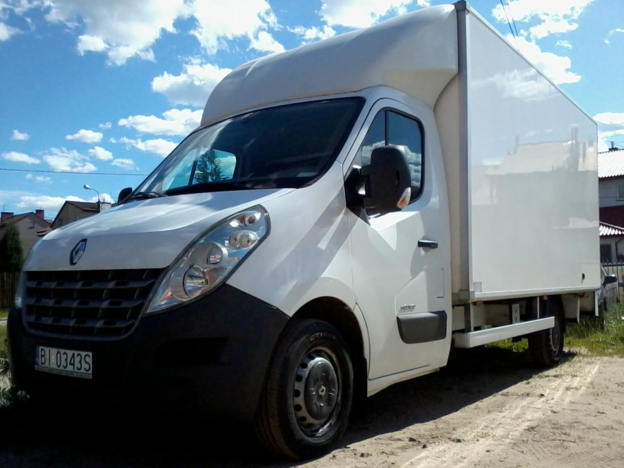 Biały samochód dostawczy Renault Master z zabudową kontenerową, zaparkowany na piaszczystym podłożu, z widoczną tablicą rejestracyjną BI 0343S i fragmentem zabudowań w tle pod błękitnym niebem...