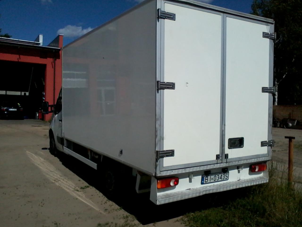 Tył białego samochodu dostawczego z zamkniętymi drzwiami, widoczna tablica rejestracyjna i fragment czerwonego budynku w tle.