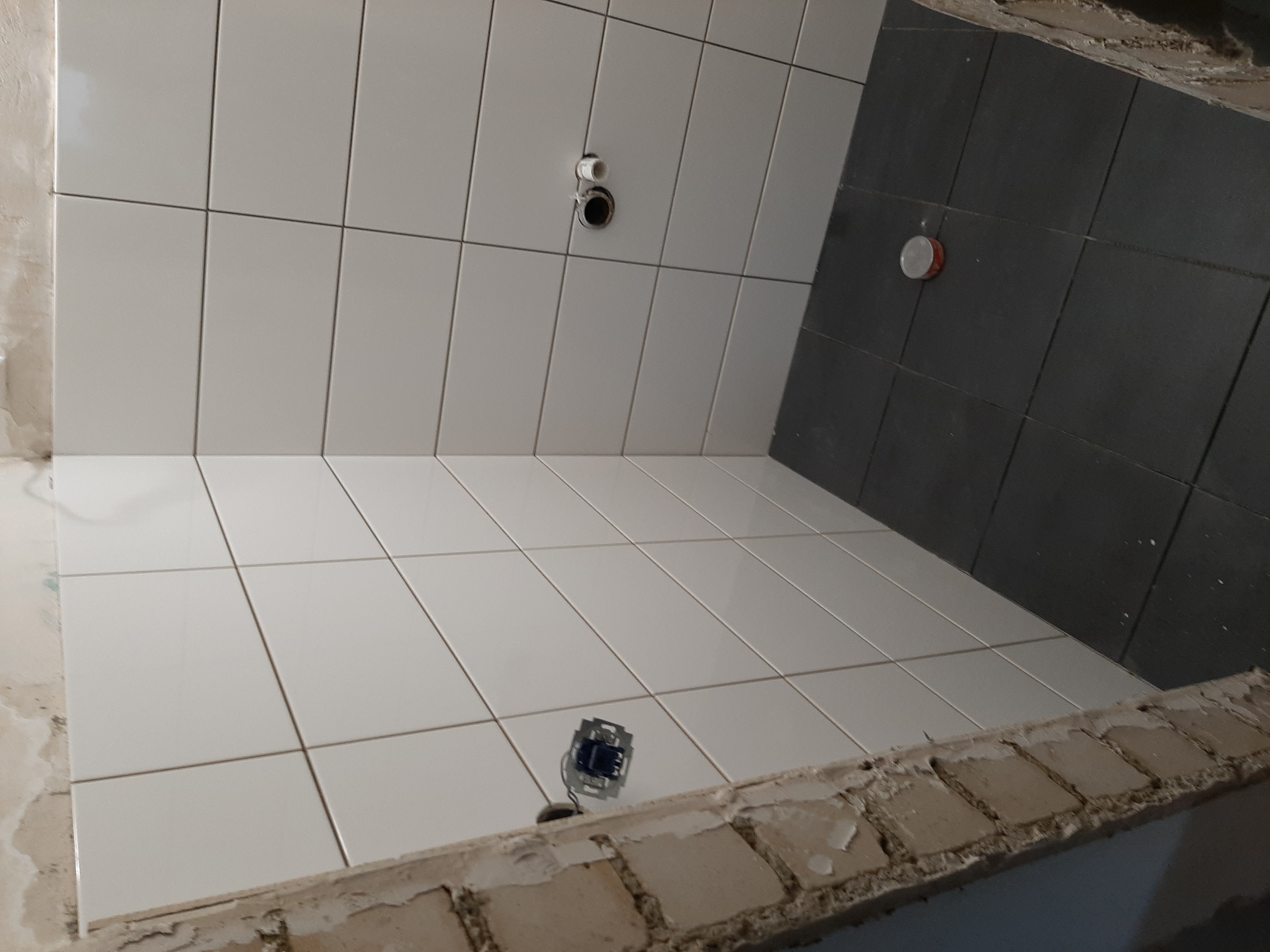 Świeżo ułożone białe i grafitowe płytki w łazience, widoczne puszki elektryczne i przyłącza wodne, stan surowy wykończenia.