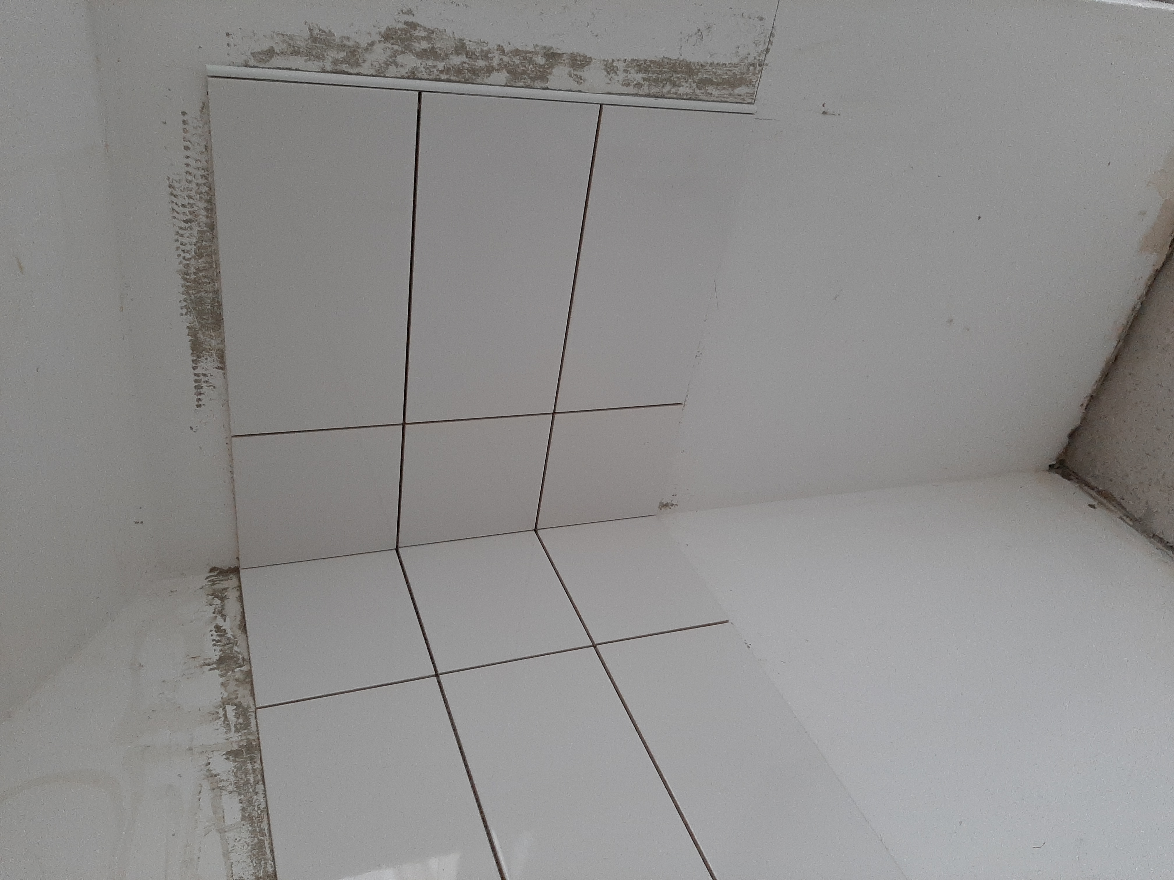 Świeżo ułożone, białe płytki ceramiczne na ścianie i podłodze w rogu pomieszczenia, widoczne ślady zaprawy na ścianach, jeszcze przed fugowaniem i ostatecznym wykończeniem.