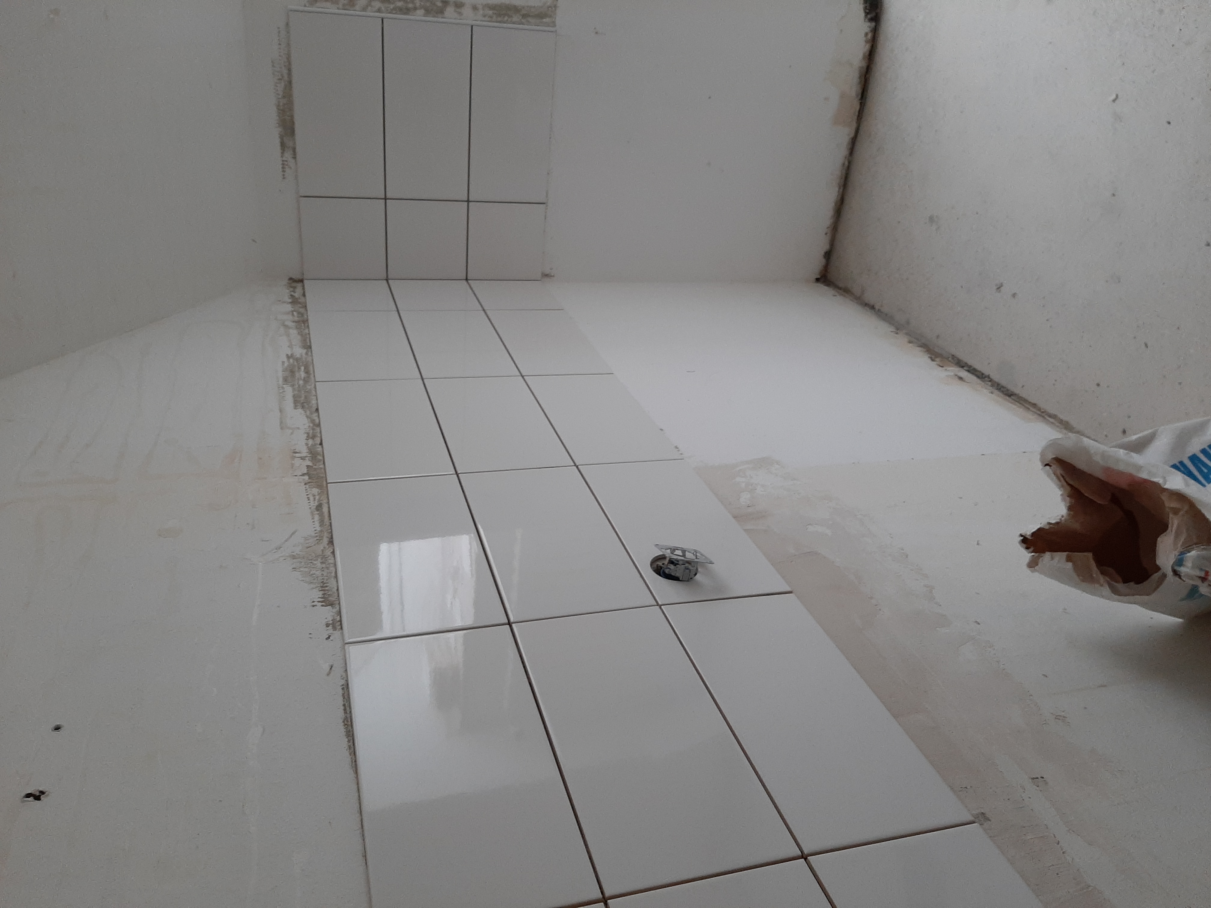 Białe, błyszczące płytki ceramiczne ułożone na podłodze i częściowo na ścianie łazienki w trakcie remontu, widoczny odpływ podłogowy i worek z zaprawą.