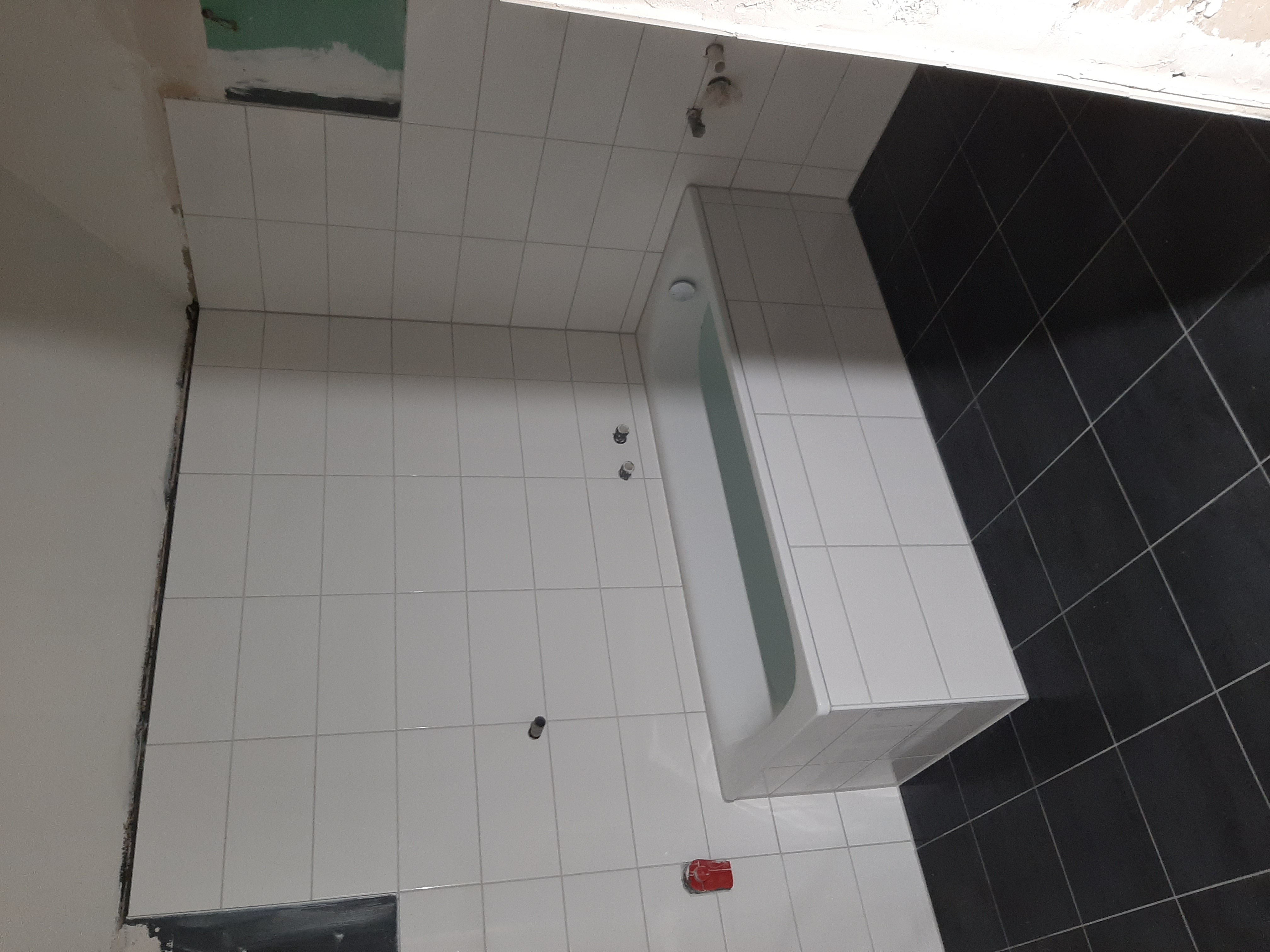 Łazienka w trakcie remontu: białe płytki na ścianach i podłodze, czarne płytki na fragmencie ściany, wanna, widoczne niedokończone prace przy suficie.