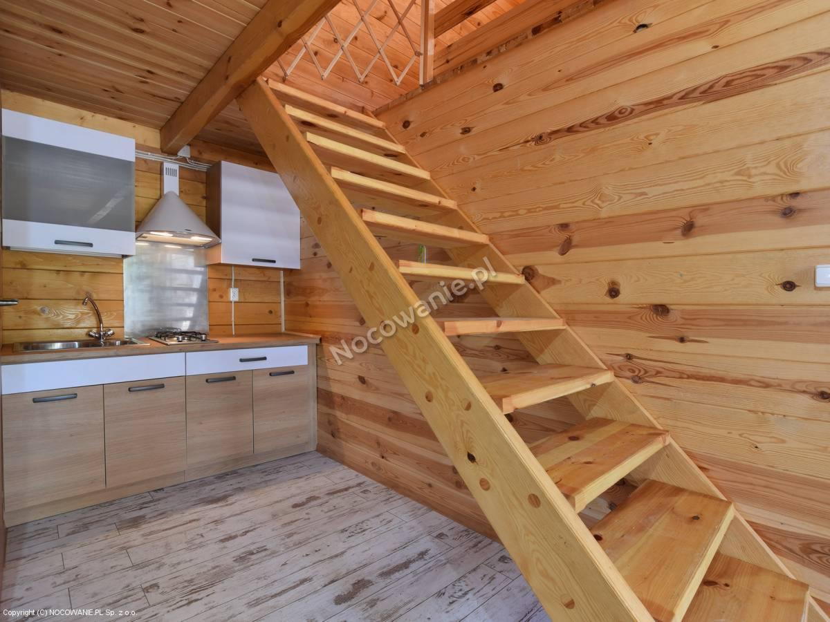 Wnętrze drewnianego domku letniskowego z aneksem kuchennym i schodami na piętro, widoczne szafki kuchenne w kolorze drewna i bieli, oraz drewniane schody.