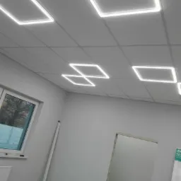 Biały sufit podwieszany z kwadratowymi panelami LED, widoczne okno i fragment ściany w szarym pomieszczeniu w trakcie prac wykończeniowych.