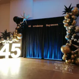 Dekoracja urodzinowa z czarnych, złotych i srebrnych balonów oraz podświetlanych cyfr '45' na tle czarnej, drapowanej ściany z napisem '45 urodziny Szymona'.