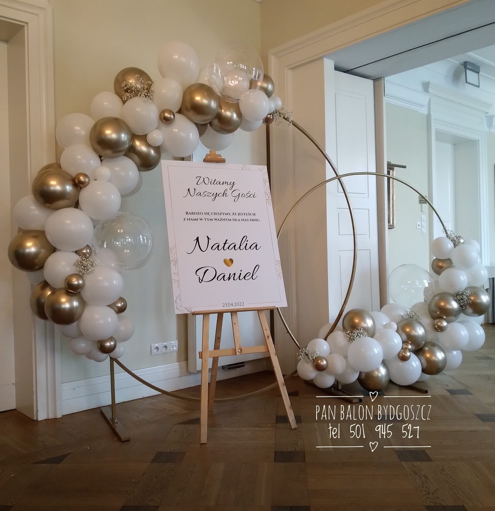 Dekoracja ślubna: złoto-białe balony ułożone w okrągłe ramy, tablica powitalna z imionami Natalii i Daniela na drewnianej sztaludze, podłoga z parkietu.