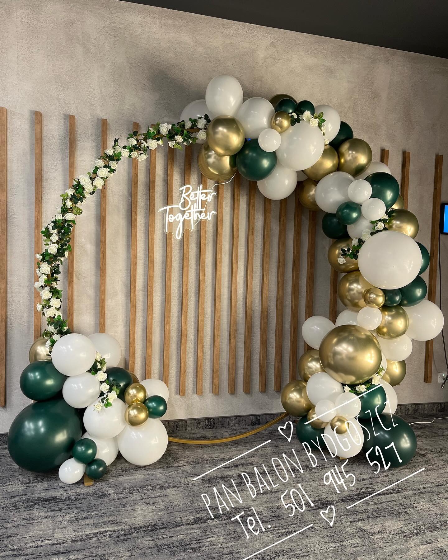 Dekoracja balonowa w kształcie łuku z kwiatami, w kolorach białym, złotym i zielonym, z neonowym napisem 'Better Together' na tle ściany z drewnianymi panelami. Na podłodze widoczny napis z numerem...