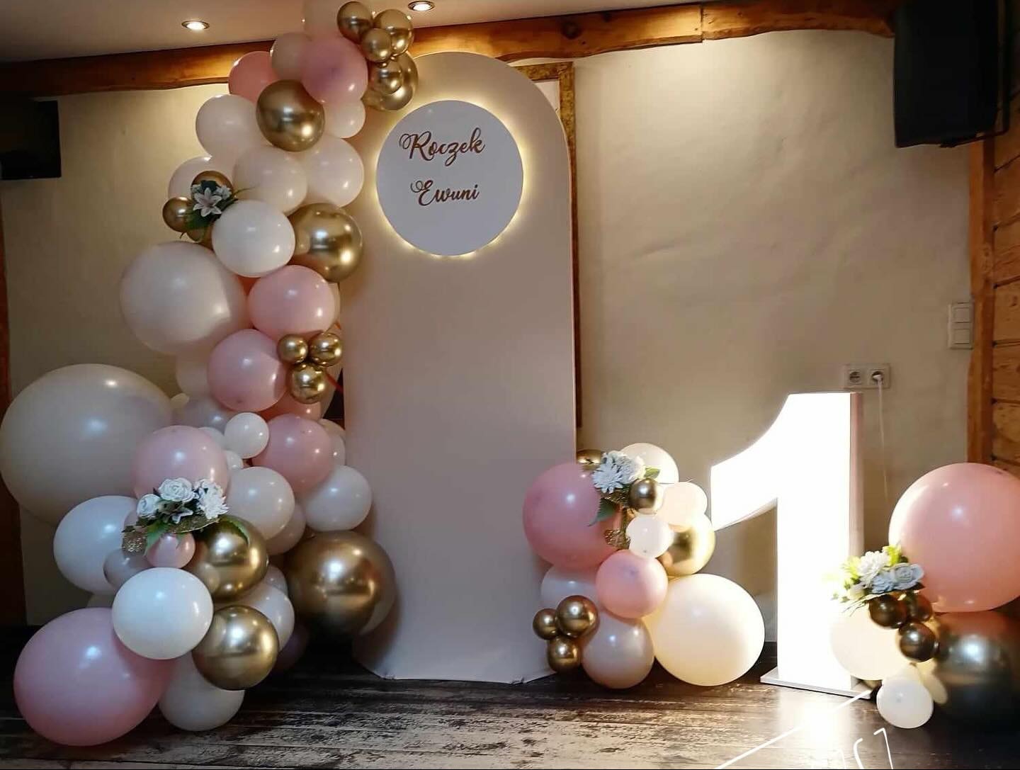Dekoracja balonowa w odcieniach różu, bieli i złota z kwiatowymi akcentami, ustawiona obok podświetlanej cyfry '1' i okrągłej tablicy z napisem 'Roczek Ewuni' na beżowym tle.