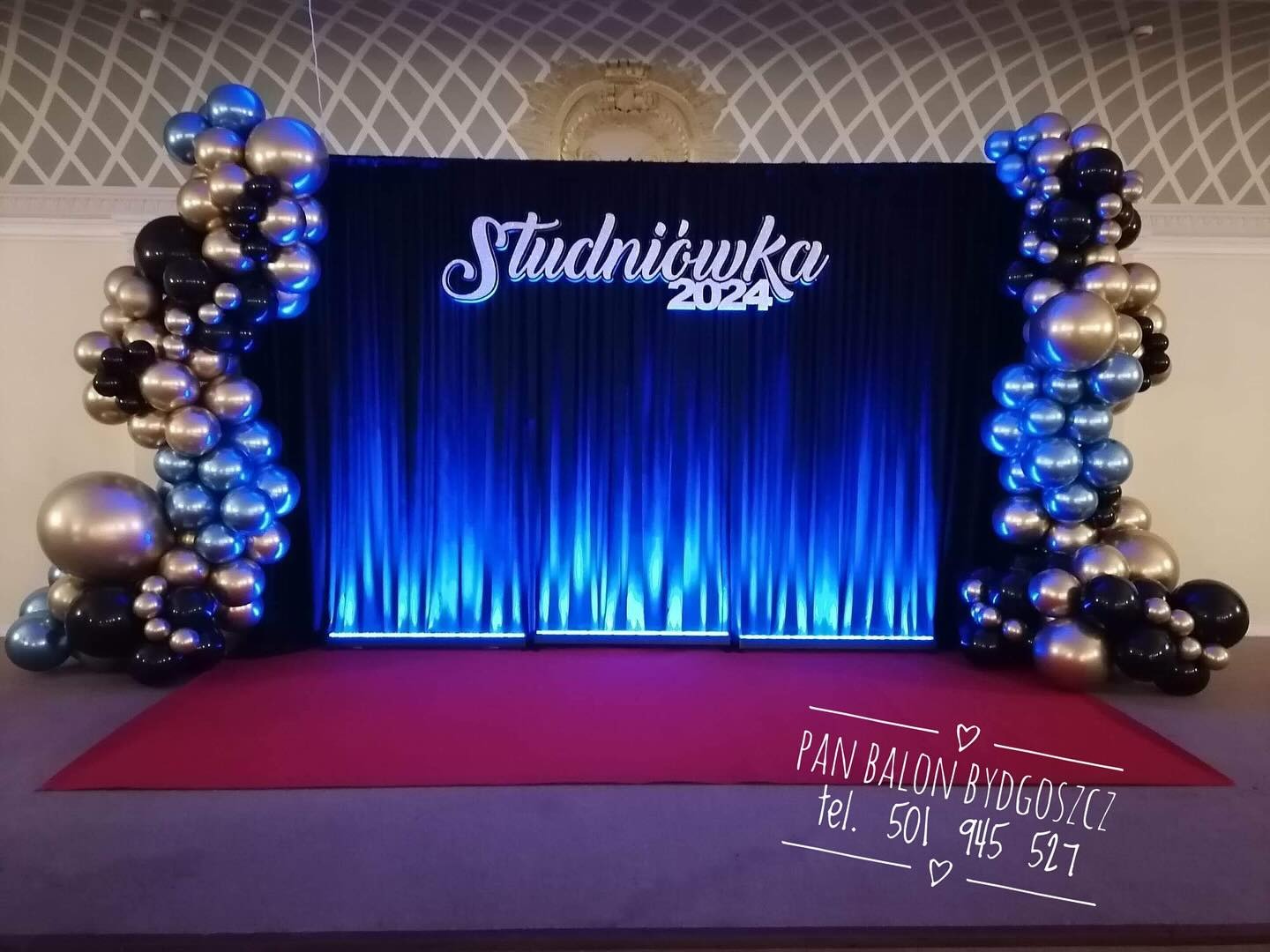 Dekoracja sceniczna na studniówkę 2024 z niebieskimi, złotymi i czarnymi balonami po bokach, podświetlona niebieskim światłem.