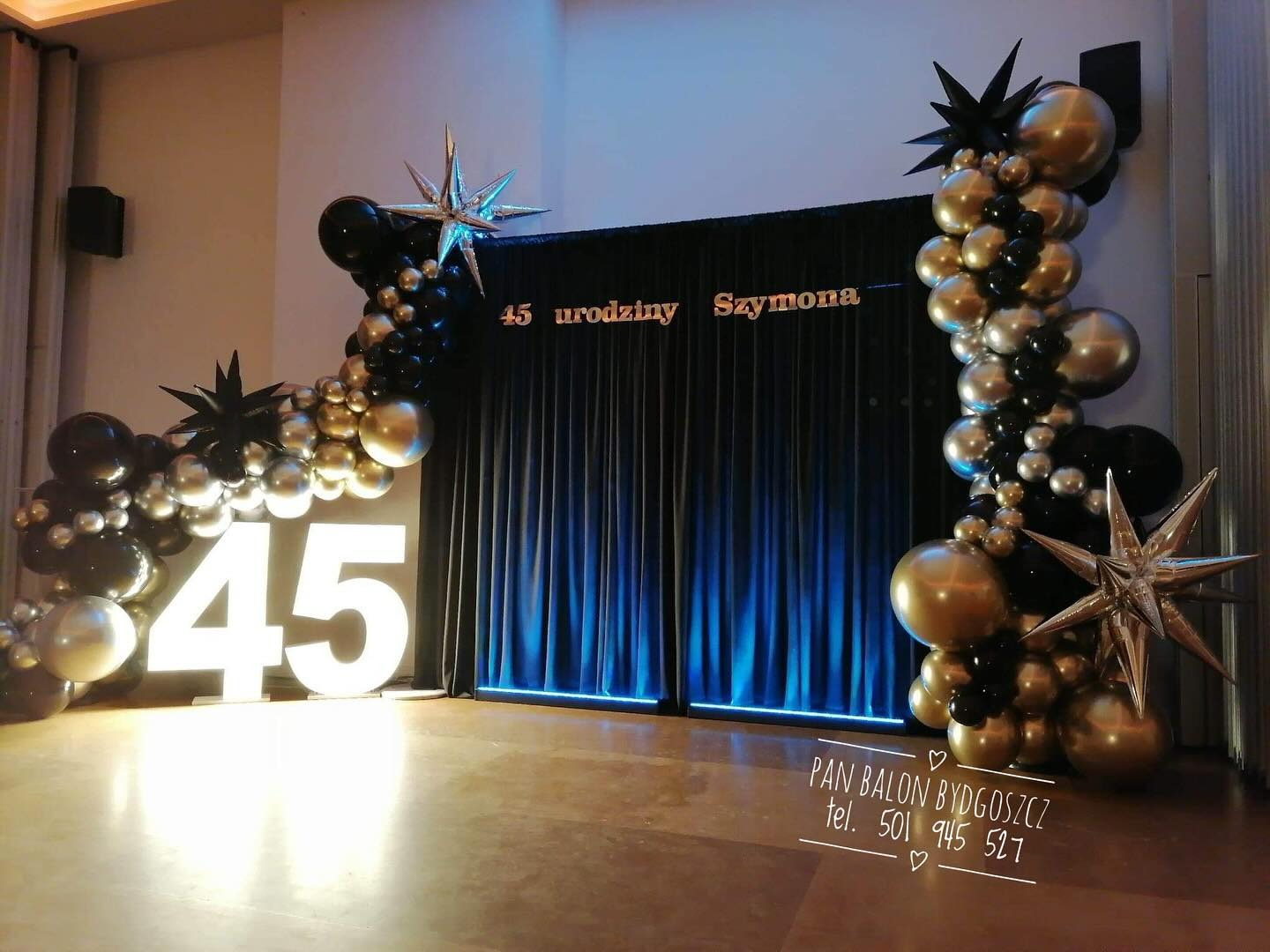 Dekoracja urodzinowa z czarnych, złotych i srebrnych balonów oraz podświetlanych cyfr '45' na tle czarnej, drapowanej ściany z napisem '45 urodziny Szymona'.