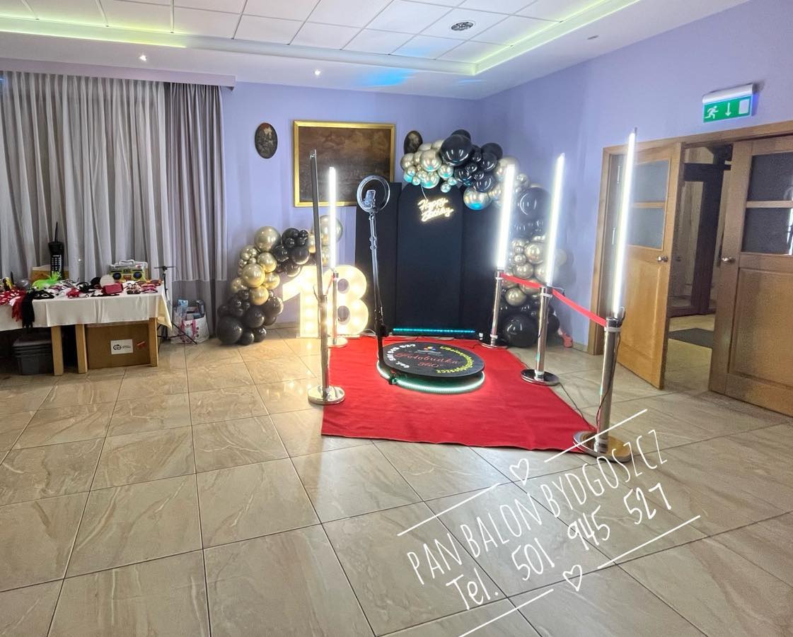 Rozstawiona fotobudka 360 na czerwonym dywanie z dekoracją balonową w odcieniach czerni i złota, oświetlona lampami ledowymi, z rekwizytami na stole w tle.