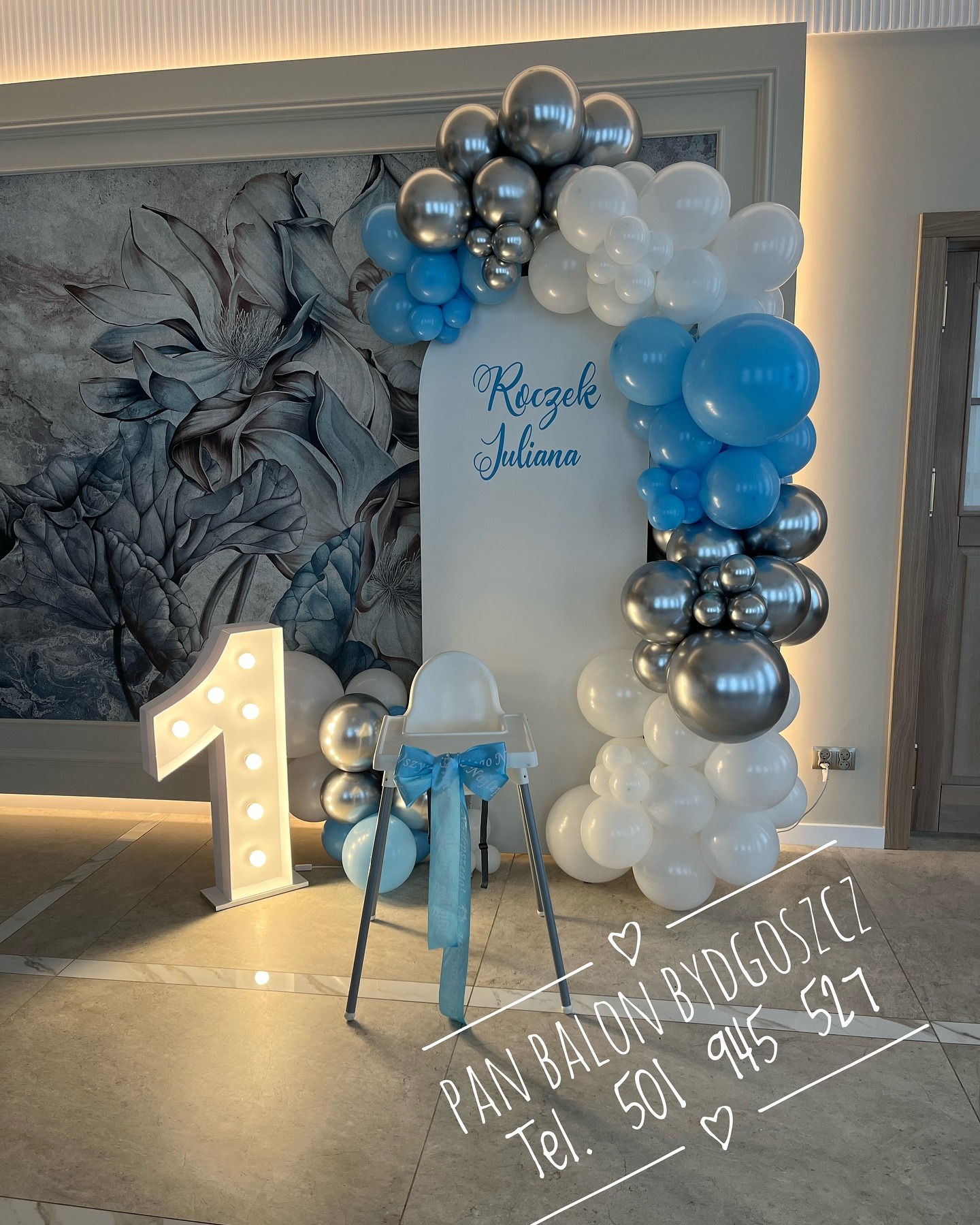 Dekoracja balonowa na roczek dziewczynki: biało-srebrno-niebieskie balony tworzące łuk, podświetlana cyfra '1', krzesełko do karmienia z niebieską kokardą, na tle ściany z motywem kwiatowym.