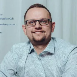 Portret uśmiechniętego mężczyzny w okularach i wzorzystej koszuli na tle jasnej ściany z tekstem.