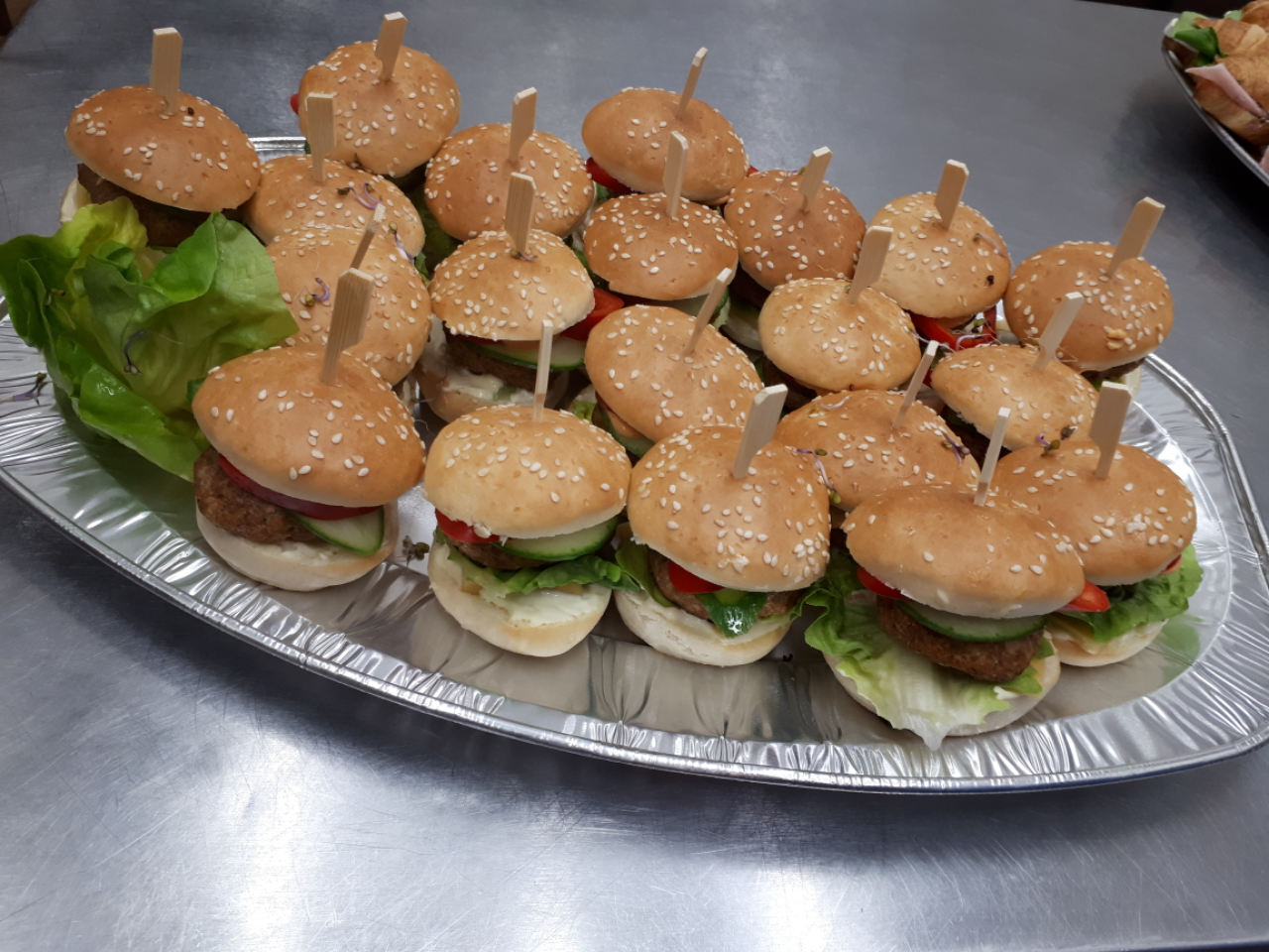 Taca z aluminiowej folii wypełniona małymi burgerami z sezamową bułką, spiętymi wykałaczkami, z dodatkiem sałaty, pomidora i ogórka.