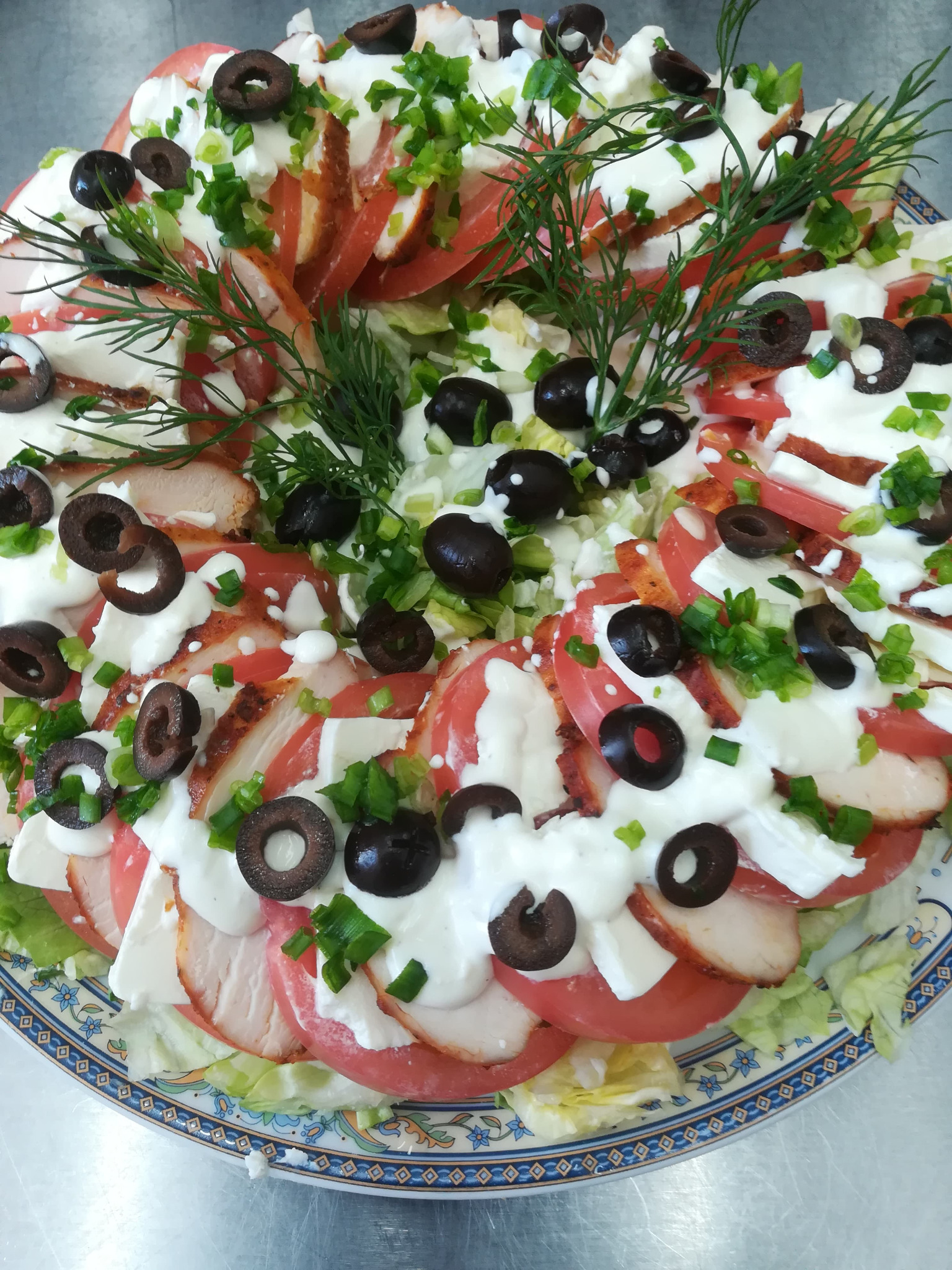 Apetyczna kompozycja warstwowa na talerzu: plasterki grillowanego kurczaka, pomidory, ser mozzarella, czarne oliwki, sos śmietanowy i posiekany szczypiorek, udekorowana gałązkami kopru.