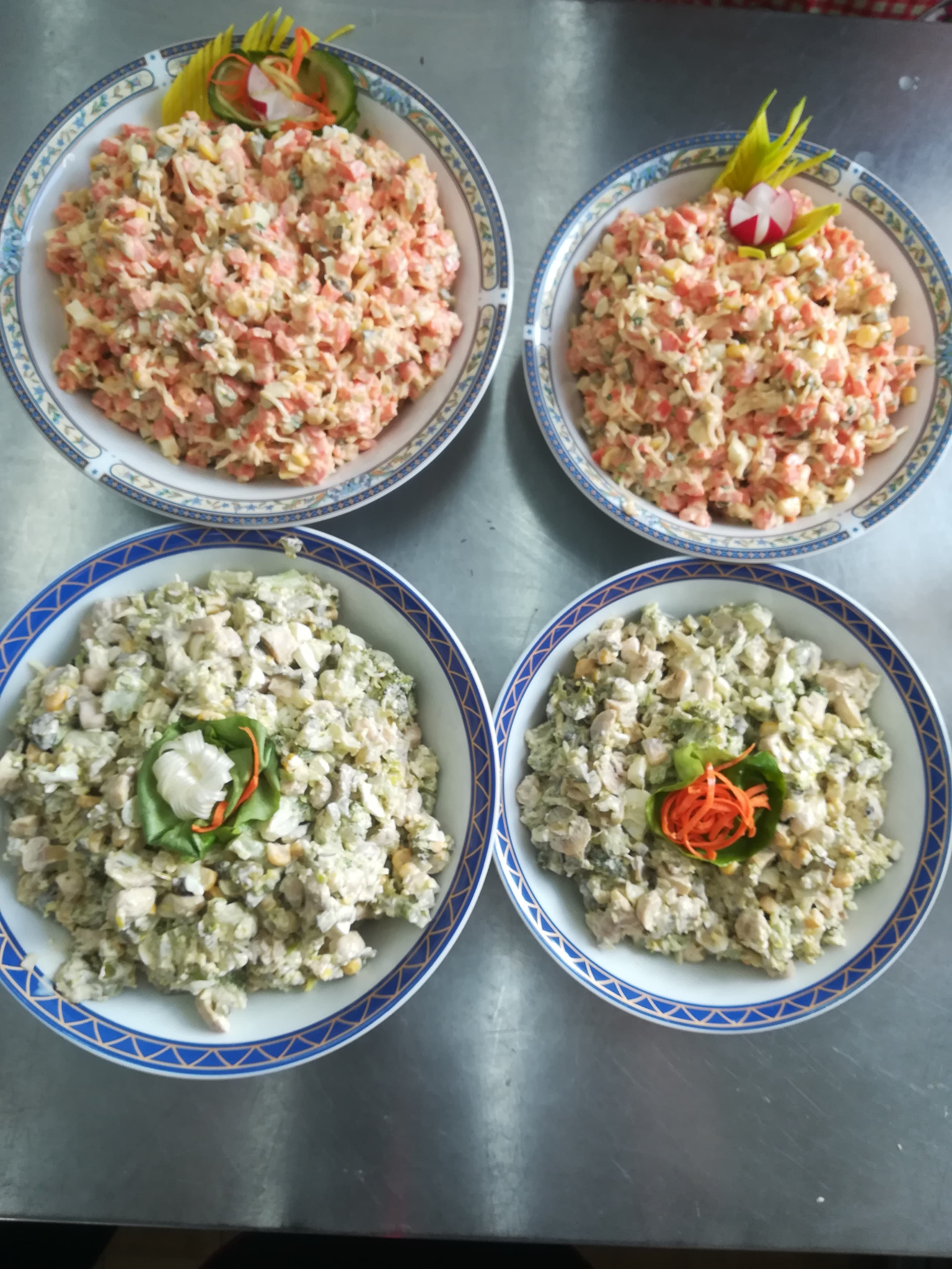 Cztery miski z sałatkami, dwie z sałatką jarzynową z dekoracją z rzodkiewki i ogórka, dwie z sałatką z kurczakiem i brokułami z dekoracją z marchewki, na metalowym blacie.