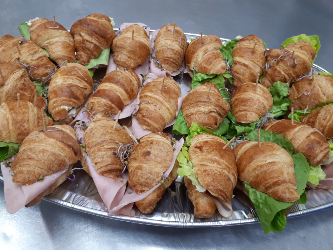 Srebrna taca z rzędami mini kanapek z croissantów, wypełnionych szynką, serem, sałatą i kiełkami, zabezpieczonych wykałaczkami.