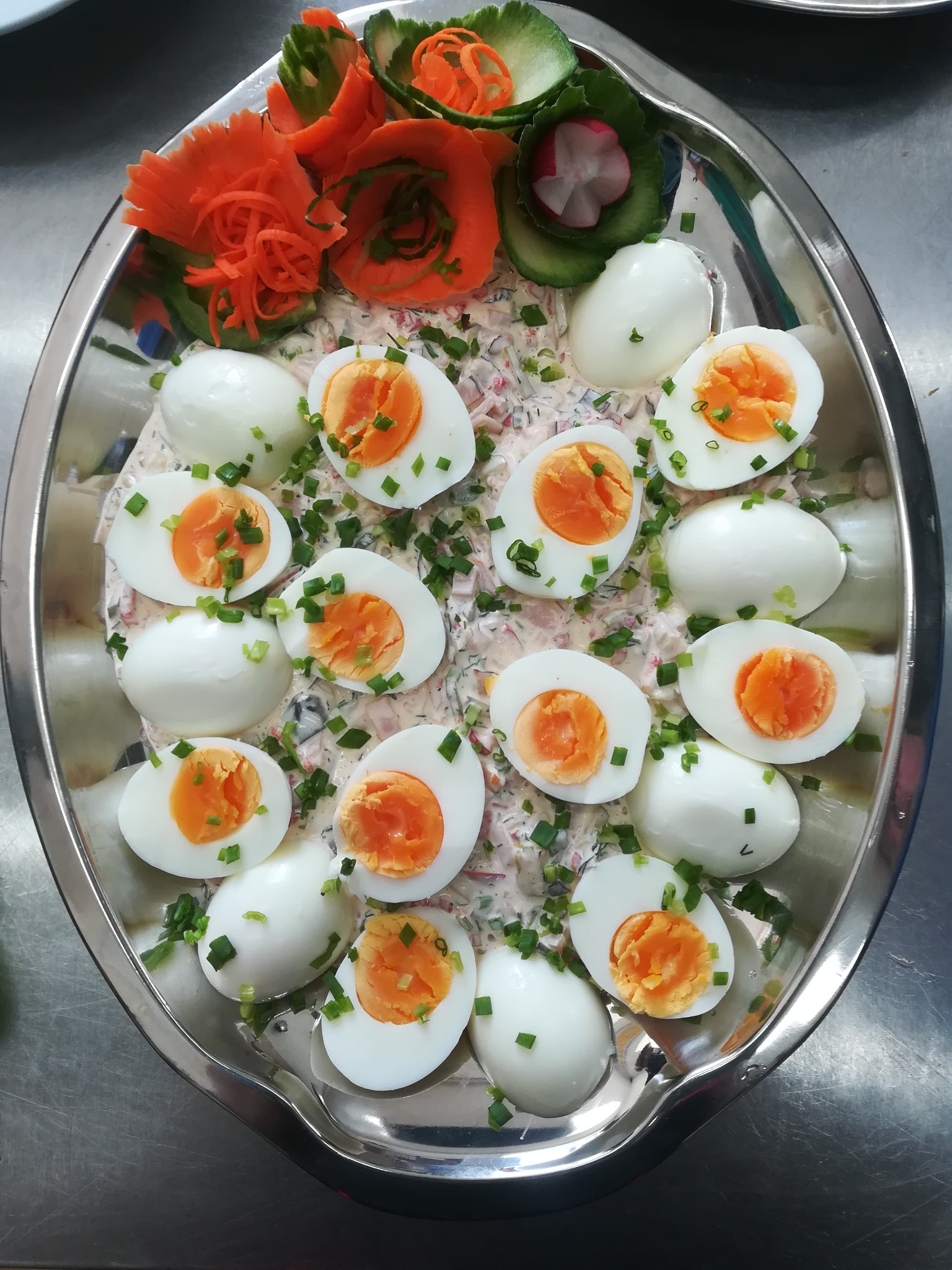 Owalna, srebrna taca z jajkami w majonezie, udekorowana rzodkiewką i marchewką wyciętą w kształt kwiatów, posypana szczypiorkiem.