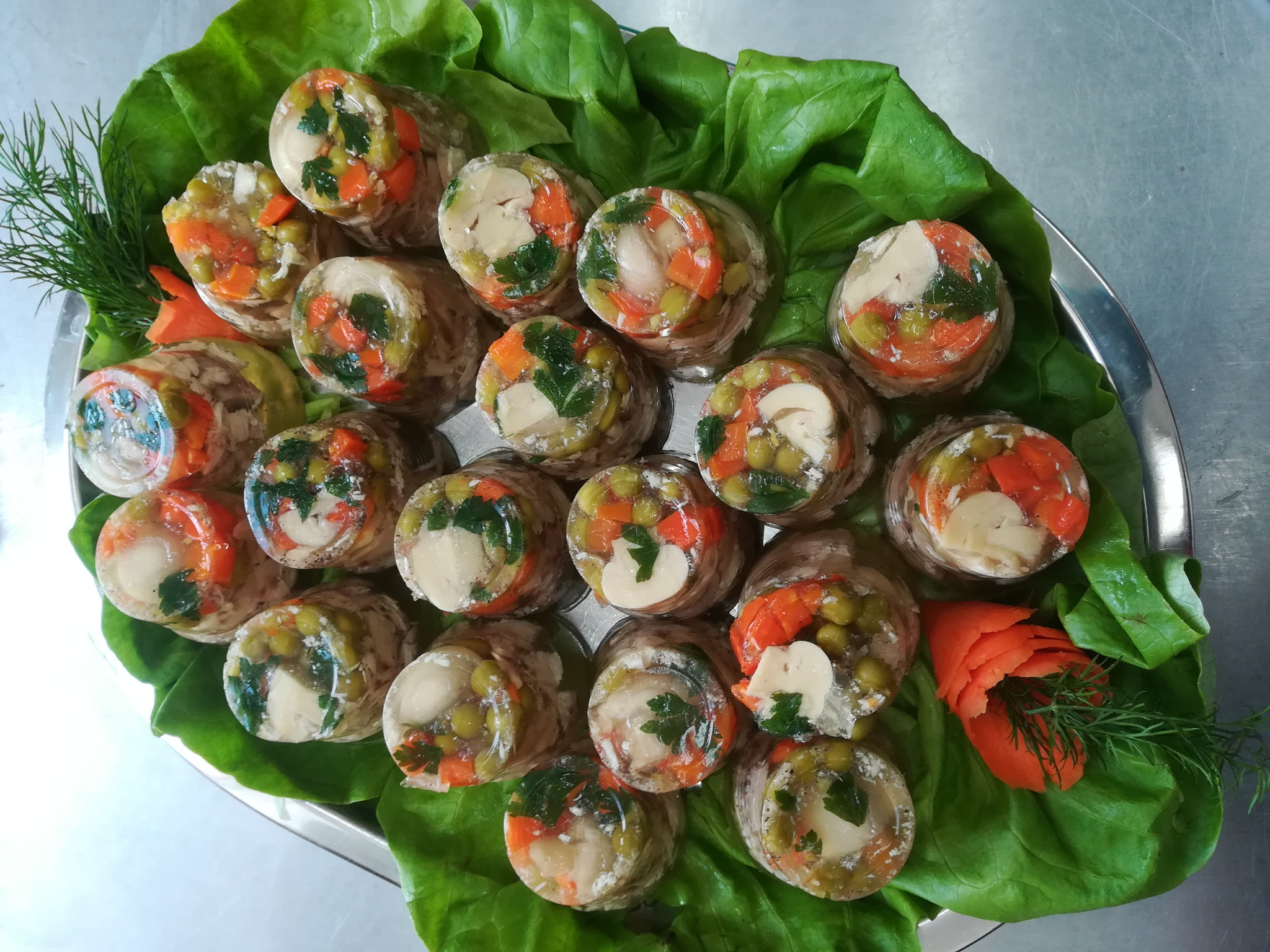 Galaretki mięsne z warzywami (marchewka, groszek, pietruszka) ułożone na srebrnej tacy wyłożonej liśćmi sałaty.