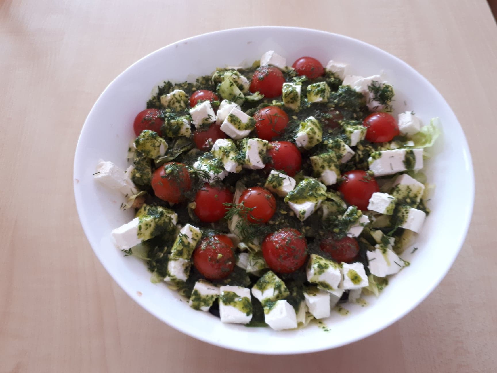 Sałatka z pomidorków koktajlowych, sera feta w kostkach i zielonego pesto, podana w białej misce na drewnianym stole.