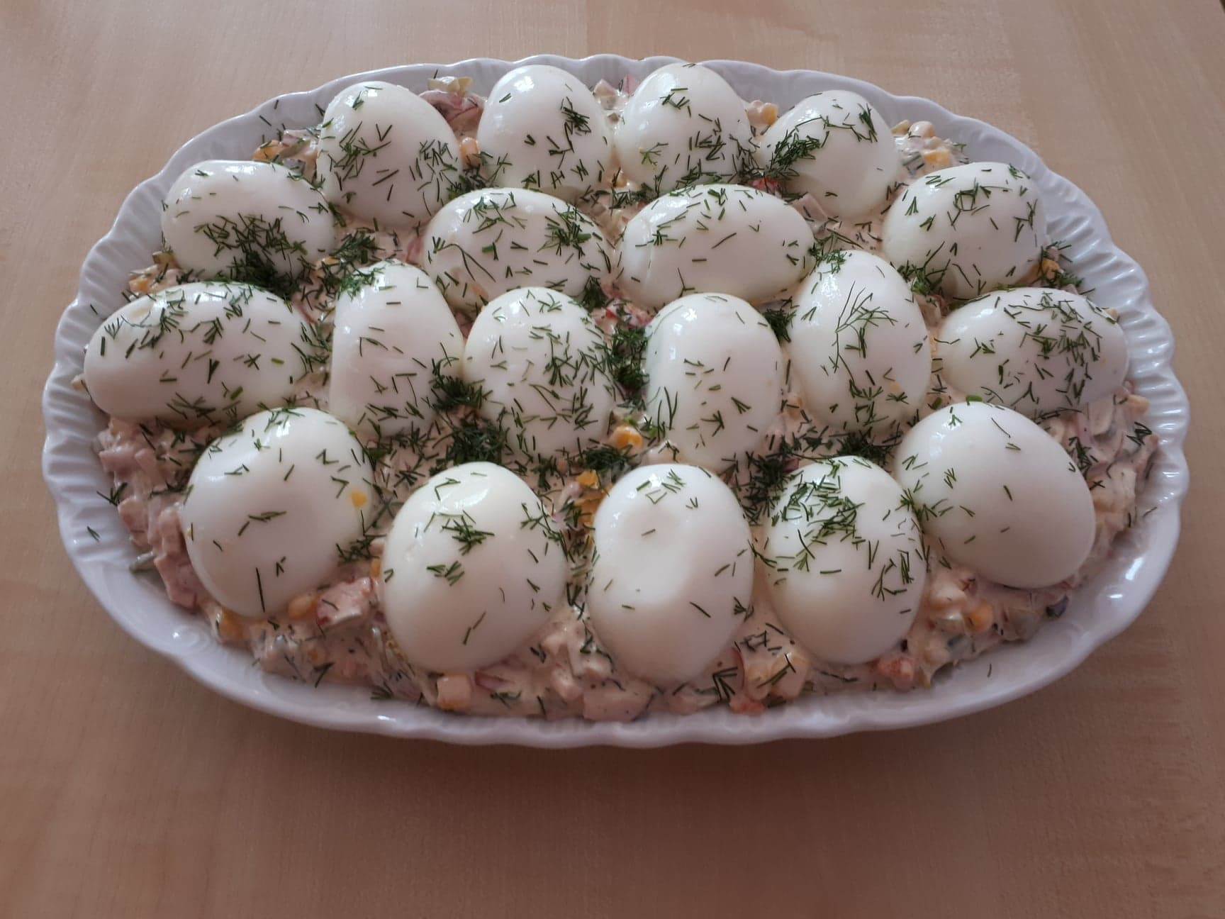 Owalny biały talerz z jajami w majonezie posypanymi koperkiem, ułożonymi na sałatce warzywnej.