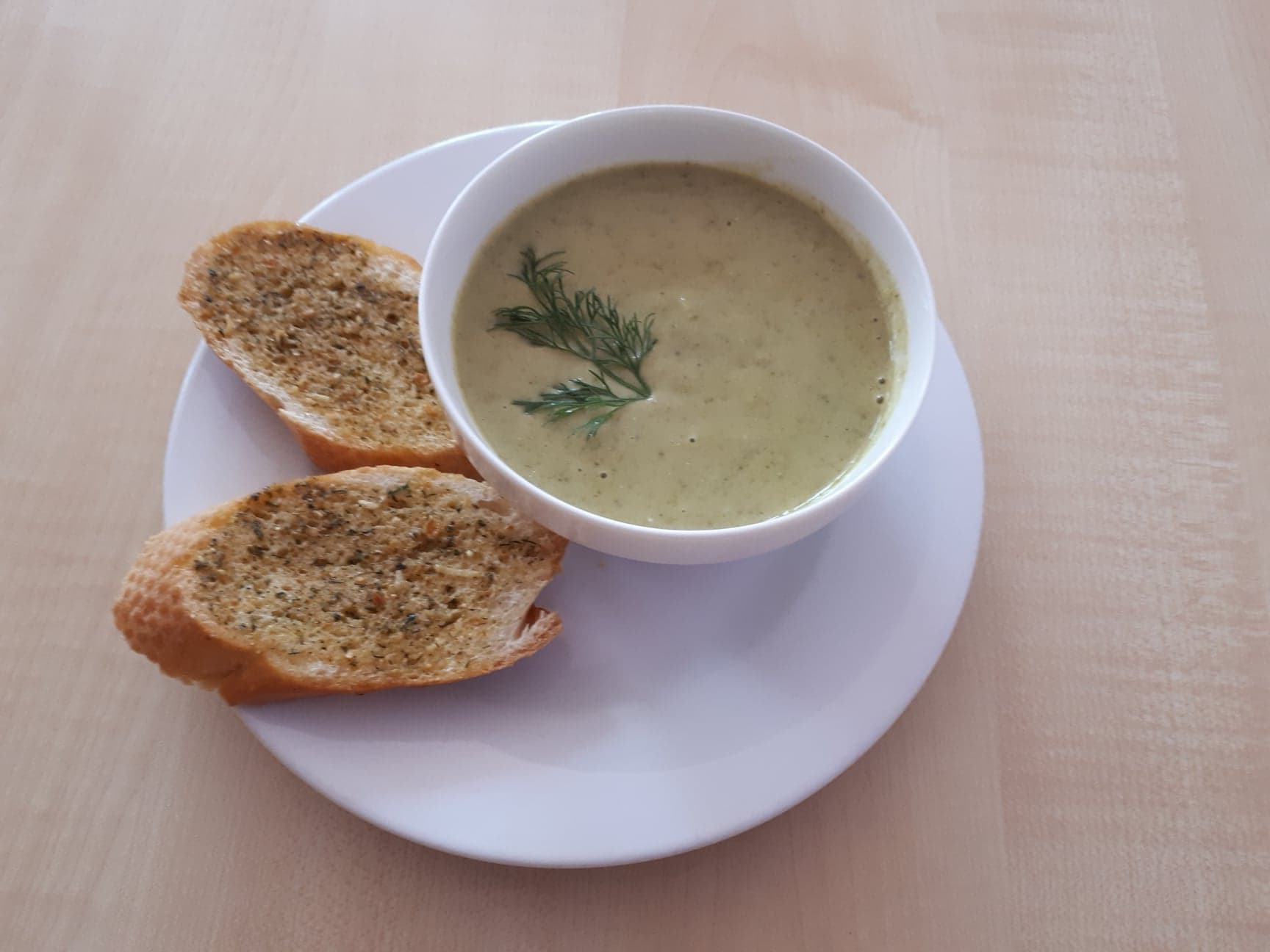 Kremowa zupa w białej miseczce, udekorowana gałązką kopru, podana z dwoma kromkami chleba czosnkowego na białym talerzu.