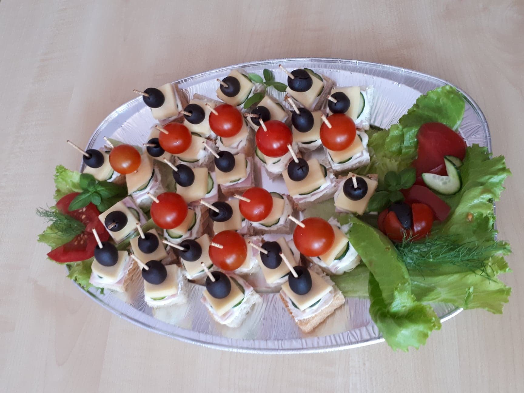 Srebrna taca z kanapkami na krakersach, z kremowym serem, szynką, ogórkiem, serem, pomidorkami cherry i czarnymi oliwkami, udekorowana sałatą, pomidorem, ogórkiem i koperkiem.