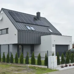Nowoczesny dom jednorodzinny z panelami fotowoltaicznymi na grafitowym dachu. Biała elewacja, numer domu 22, szara brama i ogrodzenie. W tle pochmurne niebo.