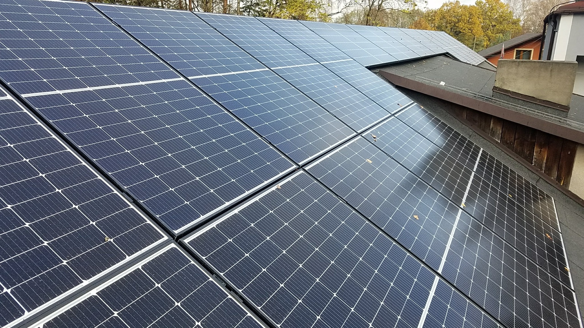 Panele słoneczne na dachu domu jednorodzinnego, widoczne opadłe liście na panelach, w tle drzewa z jesiennymi liśćmi.